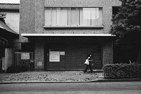 grayscale photo, woman, walking, beside, building, house, daytime, builsing, shutter, store, closed, street, urban, hedge, pavement, road, sidewalk, gate, female, bag, window, architecture, building exterior, built structure, full length, one person, real people, day, men, lifestyles, plant, leisure activity, side view, casual clothing, tree, footpath, outdoors, nature, 5K, CC0, public domain, royalty free