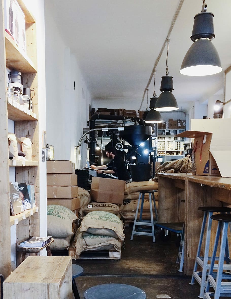 uomo, seduta, davanti, illuminato, tavolo, circondato, scatole, trasformato, grigio, lampada a sospensione, laboratorio, luci, sacchi, scaffali, contatore, in casa, nessuno, attrezzatura per l'illuminazione, mensola, tecnologia, lampada elettrica, grande gruppo di oggetti, attività commerciale, macchinario, contenitore, legno - materiale, pila, mobilia, interno di casa, giorno, natura morta, architettura, vecchio, disordinato, caffetteria, 2K