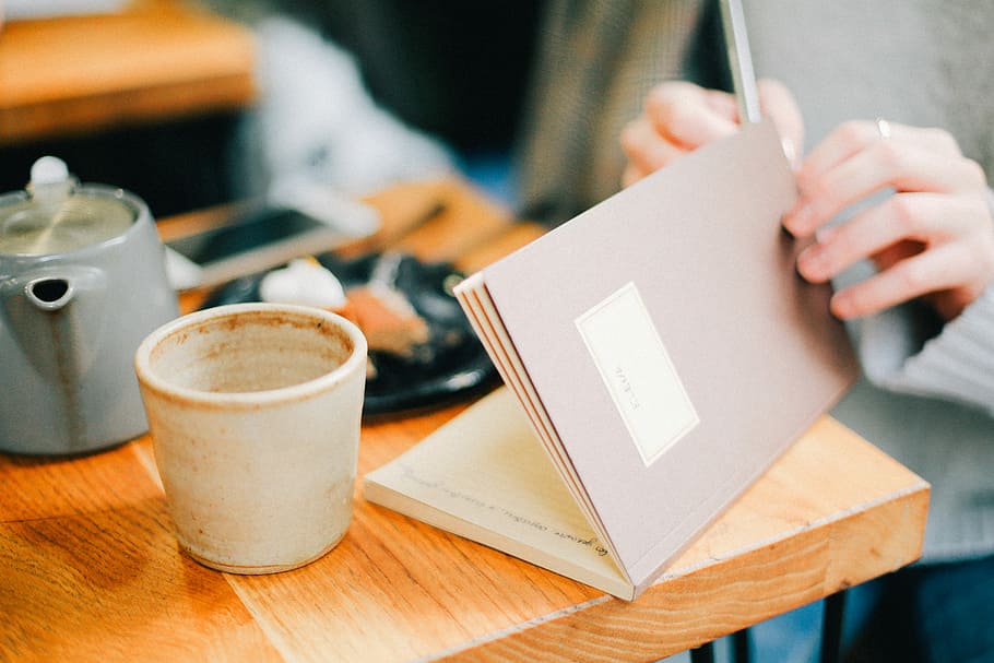 persoon openingsboek, koffiekop, persoon, bezit, boek, wit, zittend, voorkant, tafel, koffie, cafe, mok, kop, thee, een persoon, drinken, eten en drinken, binnenshuis, selectieve aandacht, koffie - drankje, focus op de voorgrond, detailopname, volwassen, menselijke hand, hand-, papier, 5K