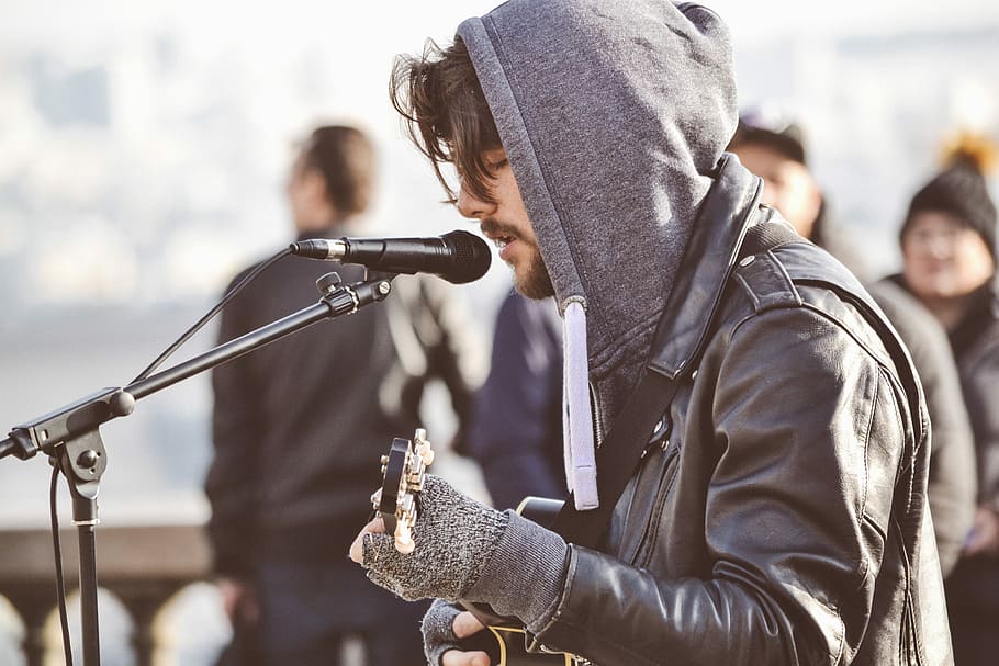 home, de cara, de costat, jugant, guitarra, durant el dia, cantar, cantant, músic, música, micròfon, bokeh, a l'aire lliure, grup, gent, dessuadora, jaqueta, fred, llum solar, ombra, ciutat, celebració, homes, campana, adult jove, caputxa - roba, arma, samarreta amb caputxa, home jove, adult, dia, de peu, roba, arquitectura, temes socials, roba casual, vida de ciutat, grup de persones, agressió, 2K