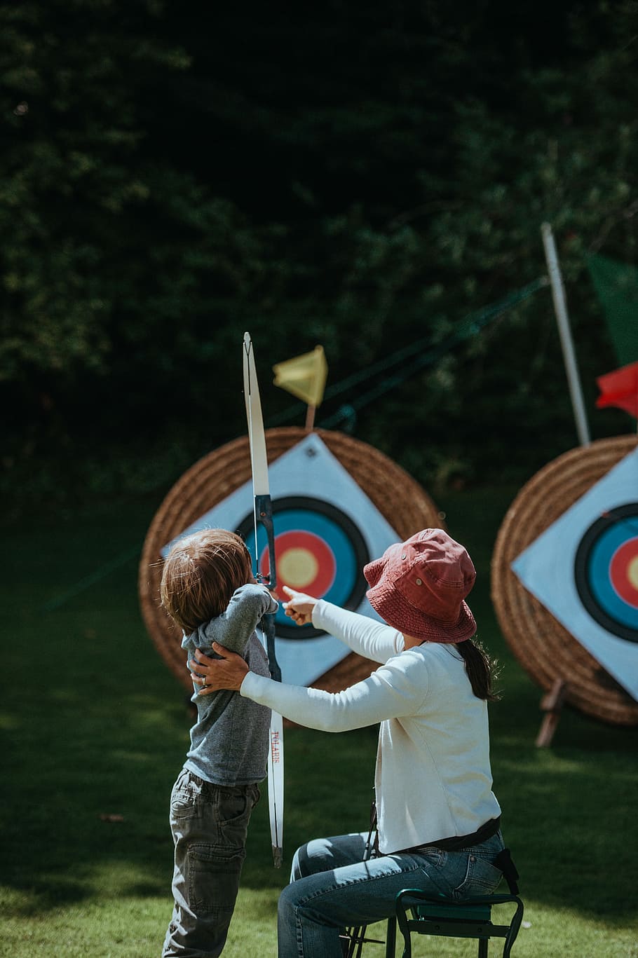 tiro con l'arco per l'apprendimento dei bambini, donna che insegna al bambino, sparare, freccia, bersaglio, arco, tiro con l'arco, ragazzo, bambino, persone reali, uomini, attività di svago, infanzia, due persone, maschi, ragazze, giorno, stili di vita, ragazzi, donne, retrovisore, femmine, solidarieta, tre quarti, abbigliamento casual, natura, persone, parco, emozione positiva, all'aperto, 4K