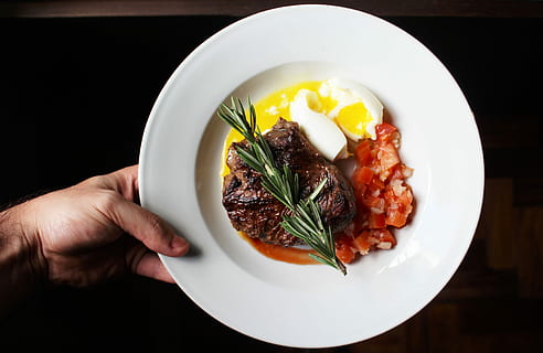 person, viser, bøf, æg, tomat, hvid, keramisk, plade, keramisk plade, mad, gourmet, måltid, aftensmad, mad og drikke, porcelæn, restaurant, kød, grøntsag, lavede mad, frokost, tæt på, madlavning, sund kost, menneskelig kropsdel, menneskelig hånd, friskhed, velvære, indendørs, hånd, klar til at spise, direkte over, en person, sort baggrund, bord, bedrift, finger, urt, pynt, 5K, CC0, offentligt domæne, royaltyfrie