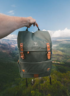 person, holding, black, brown, vinta backpack, gray, knapsack, Vinta, co, backpack, rucksack, bag, canvas bag, mountain, outdoors, nature, adventure, hiking, travel, human hand, day, one person, human body part, sky, hand, real people, environment, land, landscape, field, plant, close-up, focus on foreground, finger, 4K, CC0, public domain, royalty free
