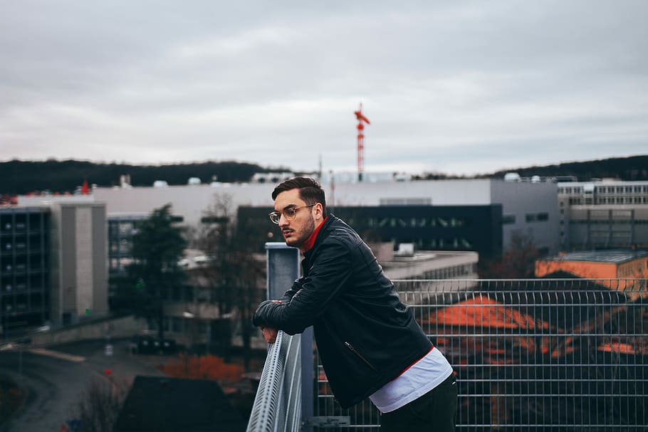 man, standing, besides, railings, leaning, gray, metal rail, portrait, jacket, glasses, cloudy, young adult, architecture, one person, sky, building exterior, built structure, cloud - sky, lifestyles, real people, young men, leisure activity, city, nature, waist up, front view, clothing, looking at camera, day, outdoors, cityscape, scarf, warm clothing, 5K