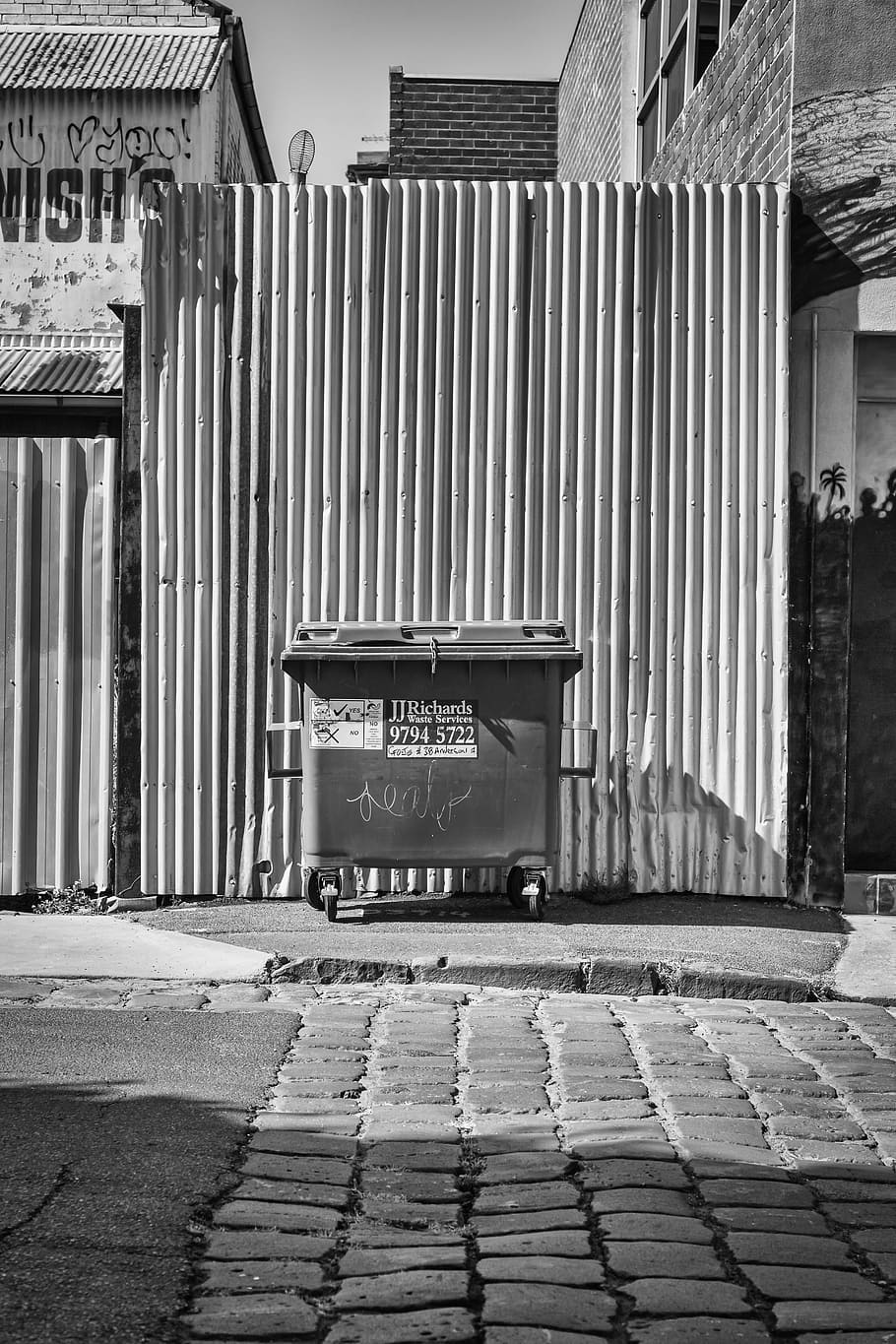 Graustufenfoto, rollen, Müll, Zaun, Behälter, Pflaster, Bürgersteig, Gebäude, die Architektur, gebaute Struktur, Gebäudehülle, Stadt, Straße, Kommunikation, Tag, Text, keine Leute, westliche Schrift, Fußweg, Sonnenlicht, Natur, draußen, Zeichen, Schatten, Metall, Eisen, gewellt, 5K