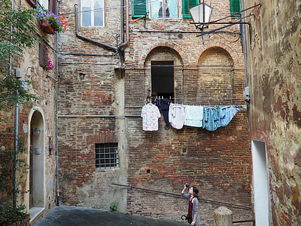 woman, gray, cardigan, holding, camera, taking, photo, floral, arrangement, 3rd, 3 rd storey window, brick, residential, building, daytime, floral arrangement, storey, window, residential building, architecture, europe, building Exterior, italy, street, built Structure, outdoors, urban Scene, old, architecture And Buildings, house, facade, brick wall, door, arch, day, residential district, wall, hanging, clothing, clothesline, no people, wall - building feature, lifestyles, 4K, CC0, public domain, royalty free