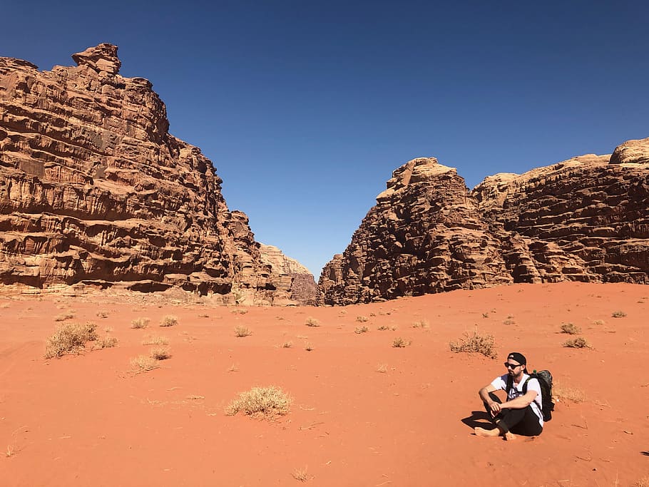 home, portar, blanc, samarreta, negre, pantalons, assegut, sòl, darrere, muntanya de formació de roca, àrid, motxilla, estèril, tapa, la llum del dia, desert, sec, geologia, herba, calenta, paisatge, muntanyes, naturalesa, a l'aire lliure, persona, roques, sorra, gres, escènic, cel, pedres, ulleres de sol, terreny erm, rock, rock - object, sòlid, escenics - natura, destinacions de viatge, viatjar, adult, formació de roca, bellesa a la natura, medi ambient, gent, escena no urbana, terra, remot, cel clar, clima àrid, clima, 4K