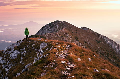 person, wearing, green, jacket, standing, cliff, facing, mountain, bubble jacket, rocky, cloudy, sky, woman, mountain top, beauty in nature, scenics - nature, sunset, tranquility, nature, tranquil scene, mountain range, leisure activity, one person, activity, non-urban scene, idyllic, orange color, environment, travel, landscape, solid, lifestyles, outdoors, mountain peak, 4K, CC0, public domain, royalty free