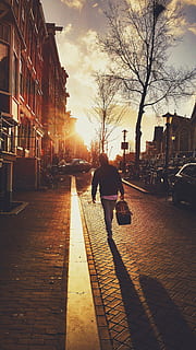 man, carrying, bag, walking, street, golden, hour, black, hoodie, sunset, cobblestone, sidewalk, pedestrian, people, duffle bag, shadow, city, streets, roads, cars, buildings, trees, sky, clouds, building exterior, architecture, city street, city life, full length, outdoors, built structure, nature, real people, one person, footpath, sunlight, tree, rear view, lifestyles, sun, lens flare, 5K