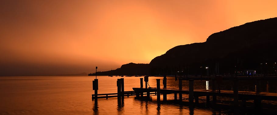 posta de sol, cinemàtic, garda, mar, naturalesa, vespre, línia de costa, aigua, escena tranquil·la, a l'aire lliure, cap gent, platja, port, estiu, cel, vacances, escenics, vaixell nàutic, imatge en color, paisatge, moll, reflexió, silueta, color taronja, bellesa a la natura, escenics - natura, tranquil·litat, muntanya, idíl·lic, terra, destinacions de viatge, cel dramàtic, cel romàntic, 5K